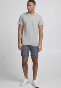 Grå polo t-skjorte med krage og tre knapper, kombinert med blå mønstrede shorts og hvite joggesko. Casual passform og glatt tekstur.