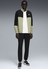 Schwarze und olivgrüne Sportjacke mit Reißverschluss, elastischen Bündchen und Logo; kombiniert mit schwarzen Hosen und schwarzen Sneakern mit weißen Akzenten.