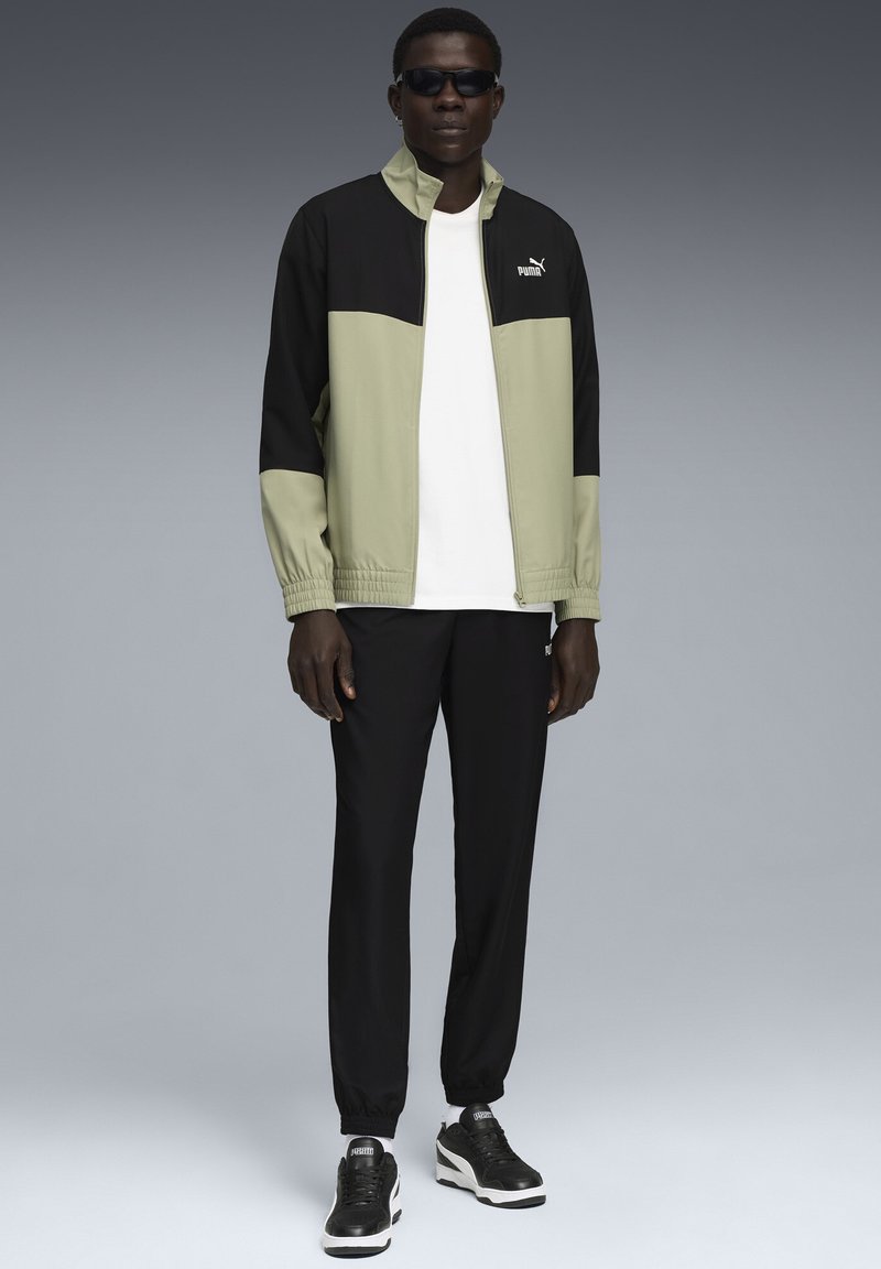 Schwarze und olivgrüne Sportjacke mit Reißverschluss, elastischen Bündchen und Logo; kombiniert mit schwarzen Hosen und schwarzen Sneakern mit weißen Akzenten.