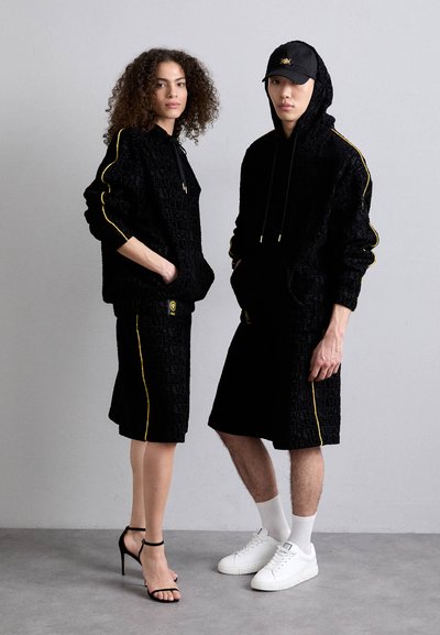 Felpa nera con texture e pantaloni short, con logo in rilievo, impreziositi da strisce gialle. Modella indossa tacchi neri; modello maschile in sneakers bianche.