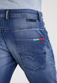Primer plano de un bolsillo de jeans de mezclilla azul con detalles bordados en rojo y verde y una etiqueta negra de la marca Diesel encima del bolsillo.