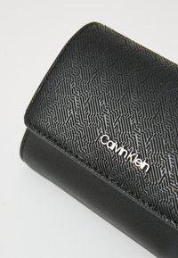 Černá peněženka s textured geometrickým vzorem a logem "Calvin Klein" v stříbrné barvě, vyrobená z syntetického materiálu, s hladkým povrchem.