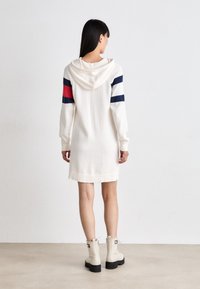Abito a maglia color crema con cappuccio e maniche lunghe, caratterizzato da strisce rosse e blu scuro sulle spalle, orlo a costine e lunghezza sopra il ginocchio. Abbinato a stivaletti beige alla caviglia.
