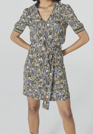 Žena v květovaných šatech se zavazovacím páskem, modrými a olivově zelenými květinami, krátkými rukávy s proužkovanými manžetami, stojící s rukama v bok.