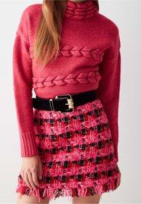Červený pletený svetr s brioškovým vzorem na límečku, kombinovaný s růžovou tkanou sukní s černým a červeným vzorem a třásněmi.