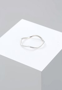 Anillo de plata ondulado con un diseño minimalista. Presenta superficies lisas y pulidas y una forma no uniforme. Exhibido en un pedestal blanco.