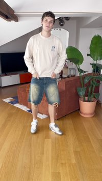 Šviesios spalvos ilgomis rankovėmis marškiniai, mėlyni džinsiniai šortai su nuskilusiomis kraštinėmis ir sidabriniai sportiniai bateliai. Stovi ant medinės grindų su augalais fone.