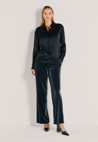 Schwarze Satinbluse mit einer Krawatten-Halsöffnung, kombiniert mit dunkelblauen, samtigen Weitbeinhosen und spitz zulaufenden schwarzen Absätzen. Glatte Textur insgesamt.