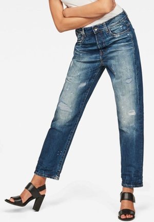 Person trägt abgenutzte blaue Jeans und schwarze Sandalen mit hohen Absätzen, steht mit gespreizten Beinen und verschränkten Armen vor der Brust.