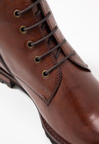 Botte en cuir marron avec œillets en métal, lacets ton sur ton et accents cousus. Présente une surface légèrement texturée et une semelle robuste.