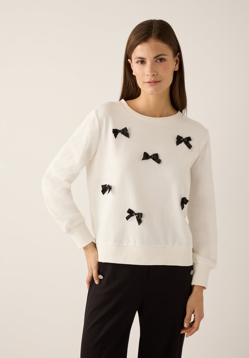 White sweatshirt with long sleeves, featuring multiple small black bows attached to the front. Ribbed cuffs and hem. Paired with black pants.