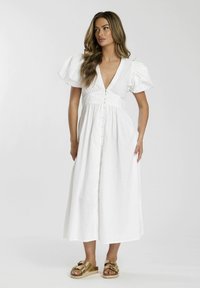 Wei−les, knielanges Kleid mit flatternden Ärmeln, tiefem V-Ausschnitt und Knöpfen auf der Vorderseite. Das Modell kombiniert es mit goldenen Sandalen. Glatte Textur.