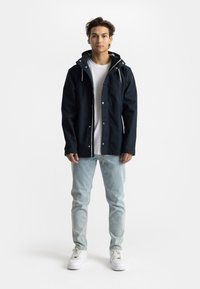 Navy waterproof jacket with drawstring hood, button closures, and zip pocket, worn over a white t-shirt and paired with light blue jeans and white sneakers.