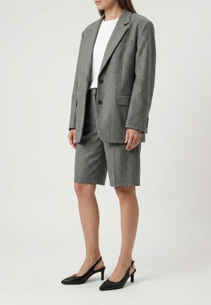 Vrouw draagt een grijze oversized blazer en bijpassende knielange korte broek, een wit topje en zwarte slingback hakken, staand tegen een effen achtergrond.