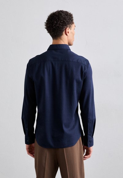 Camisa de manga larga azul marino con cuello puntiagudo, puños con botón y una textura sutil. Presenta un dobladillo recto y un detalle en el yugo trasero.