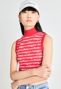 Haut sans manches rouge avec un col montant portant le texte "tommy jeans" en blanc répété. Le mannequin porte une casquette blanche et un short en denim.