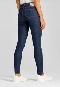 Osoba v tmavě modrých slim kalhotách a bílých slip-on botách, stojící zády k divákovi na jednobarevném světle šedém pozadí.