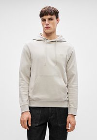 Huppari vaaleanbeige sävyssä, valmistettu pehmeästä kankaasta. Sen etupuolella on tasku, kiristettävä huppu ja pieni logopatch rintataskussa.