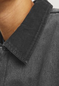 Collare in corduroy nero con righe prominenti, attaccato a un tessuto di denim scuro, che mostra una texture liscia e una leggera lucentezza.