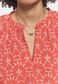 Blusa de coral con bordado floral blanco, escote en V y hombros plisados. Complementada con un sencillo collar de cadena dorada.