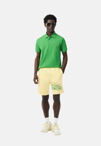 Camisa polo verde, pantalones cortos amarillos con logo verde, zapatillas blancas y gafas de sol. Diseño casual de mezcla de algodón, entallado, de manga corta.