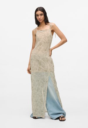 Ženska v bež cvetlični obleki brez rokavov čez široke svetlomodre kavbojke in črne sandale, stoji ob belem ozadju.