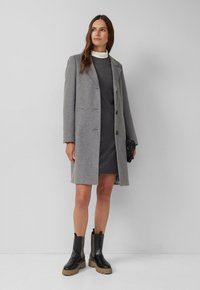 Eine Frau in einem knielangen grauen Mantel, einem dunkelgrauen Kleid, schwarzen klobigen Stiefeln, die eine kleine schwarze Handtasche hält, steht an einer schlichten Wand.