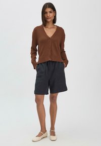 Brauner Button-up Cardigan mit V-Ausschnitt, kombiniert mit dunklen Shorts mit zwei Eingrifftaschen. Das Model trägt beige Ballerinas.