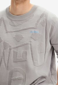 Szary sweter dziany z teksturowanymi wzorami i subtelnym niebieskim logo. Okrągły dekolt i lekka tkanina z różnorodnymi detalami szwów.