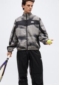 Νεαρός άνδρας που κρατάει μπάλα του τένις και ρακέτα, φορώντας αθλητικό μπουφάν με μαύρες και γκρι ρίγες και μαύρο παντελόνι, με λευκό φόντο.