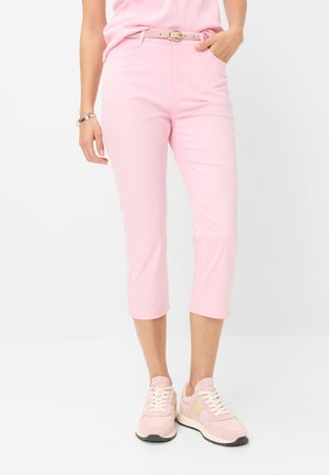 Vrouw draagt lichtroze cropped broek, bijpassende riem en sneakers, staat met één hand in de zak tegen een effen witte achtergrond.