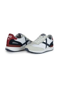 Zapatillas blancas con materiales de ante y malla, acentos en azul marino y detalle en rojo en el talón. Presentan una suela texturizada y un diseño de cordones.
