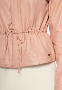 Giubbotto in pelle rosa pallido con vita cinchata e cordoncino, dettagli in hardware dorato vicino al fondo. Texture liscia.