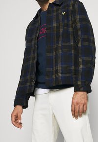 Man i en marinblå och svart rutmönstrad zip-up jacka över en marinblå sweatshirt och vita byxor, stående mot en enfärgad bakgrund.