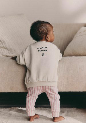 Bebeluş stând desculţ, ţinând o canapea bej, purtând un pulover crem cu textul „poveşti creative” şi pantaloni roz cu dungi.