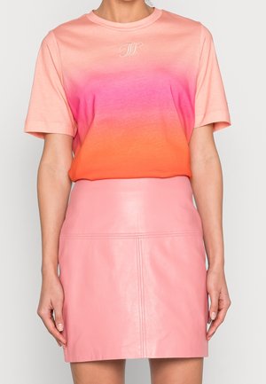 Nainen, jolla on päällään vaaleanpunaisen ja oranssin sävyinen lyhythihainen t-paita, joka on sullottu korkeavyötäröiseen vaaleanpunaiseen nahkaiseen minihameeseen, yksiväristä taustaa vasten.