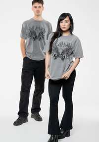 Gray graphic t-shirts with a skull and wing design, paired with black flared pants. Both models wear black shoes. Smooth fabric texture.