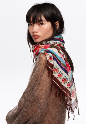 Joven con cabello largo y oscuro que lleva un suéter marrón peludo y una bufanda colorida con estampado floral sobre los hombros, mirando de lado.