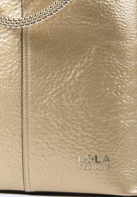 Zlatá metalická taška s textúrovaným povrchom, ktorá má striebornú reťazovú rukoväť a embosované logo na spodnej strane.