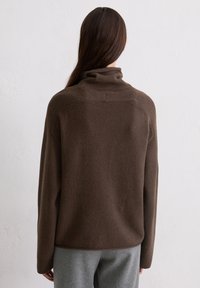 Brązowy, oversize sweter z wysokim kołnierzem o miękkiej fakturze, z rękawami raglanowymi, noszony na szarych spodniach.
