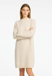 Beige stickad klänning med hög hals, långa ärmar och en avslappnad passform. Mjuk, fluffig textur med fransar vid nederkanten.