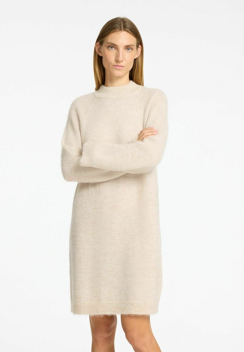 Robe en tricot beige avec un col haut, des manches longues et une coupe décontractée. Texture douce et pelucheuse avec des franges à l'ourlet.