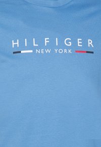Modrá látka s bílým textem "HILFIGER NEW YORK" a dvěma vodorovnými pruhy v černé, bílé, červené a černé na obou stranách "NEW YORK".