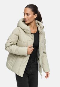 Giubbino imbottito beige chiaro con un grande cappuccio, texture trapuntata, zip frontale e tasche laterali, indossato sopra una maglietta nera e pantaloni neri.