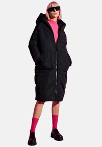 Cappotto puffo nero con cappuccio, chiusura a zip e ampie tasche anteriori; indossato sopra un maglione rosa brillante, abbinato a calzini fucsia e scarpe nere.