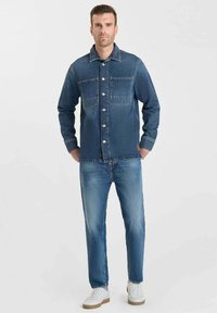 Denimjas met twee borstzakken, blauwe kleur, knoopsluiting, gedragen over lichtblauwe jeans en witte sneakers met zandkleurige zolen.