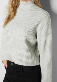 Ljusgrå ribbad stickad tröja med en hög, åtsittande halsringning och sjunkna axlar, tillsammans med svarta byxor med en metallknapp.