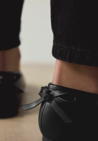 Melni ādas apavi ar apaļu purngalu un dekoratīvu loku aizmugurē, kopā ar tumša džinsa biksēm, kuras izceļas ar teksturētu audumu.