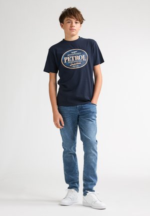 Tricou albastru marin cu un grafic alb și portocaliu „PETROL”; purtat cu blugi deschis la culoare și adidași albi. Material din bumbac fin, croială casual.