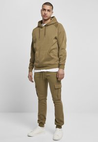 Felpa con cappuccio verde oliva con tasca frontale; abbinata a pantaloni jogger coordinati con tasche cargo e strisce laterali. Sneakers bianche.